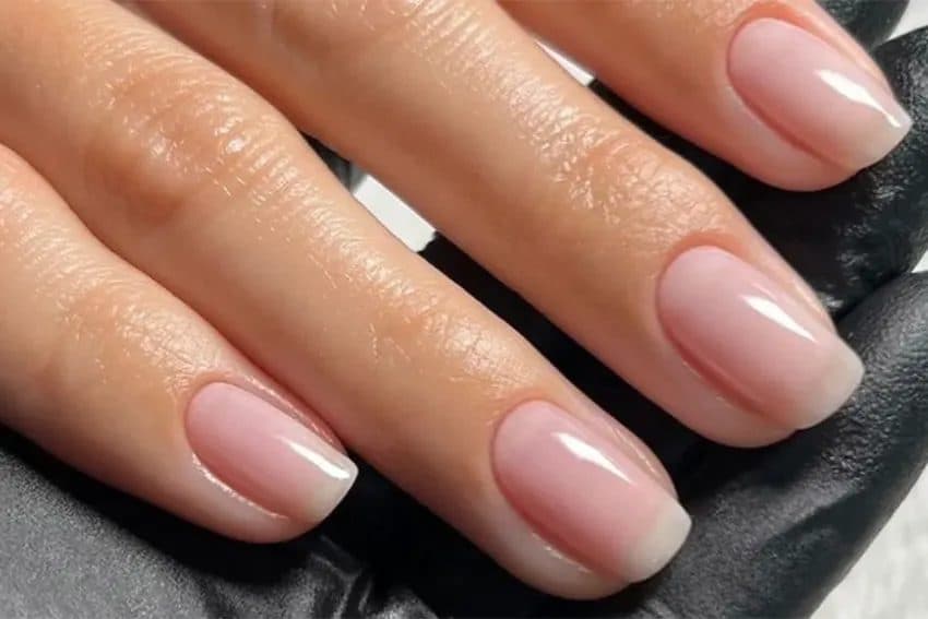 Ongles courts à la finition rose laiteux brillante, cuticules nettes, doigts posés sur une main gantée de noir.