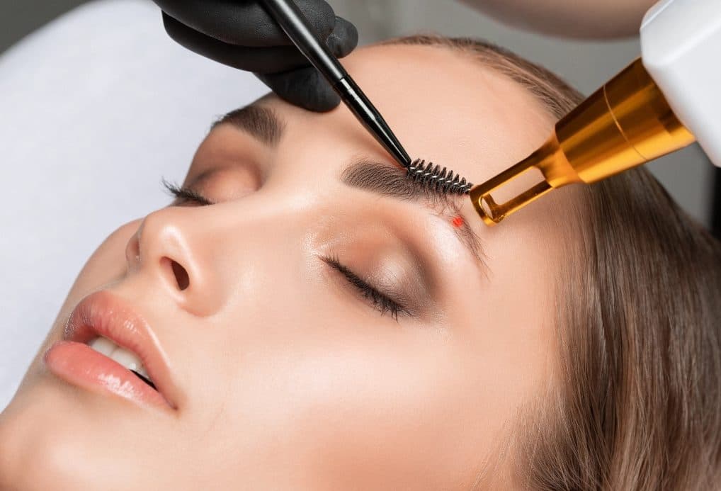 Pièce à main laser dorée pointée près du sourcil d’une cliente allongée, point de visée rouge sur la peau, poils maintenus à la brosse par une main gantée de noir.