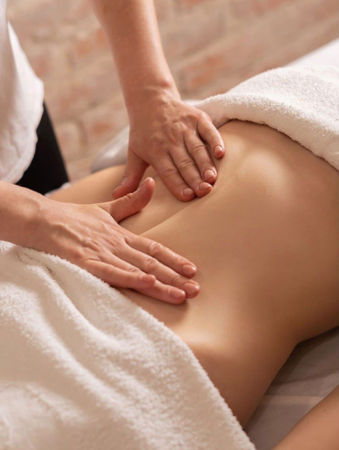 Deux mains posées à plat sur l’abdomen d’une cliente allongée entre deux serviettes éponge, pendant un soin manuel du corps.
