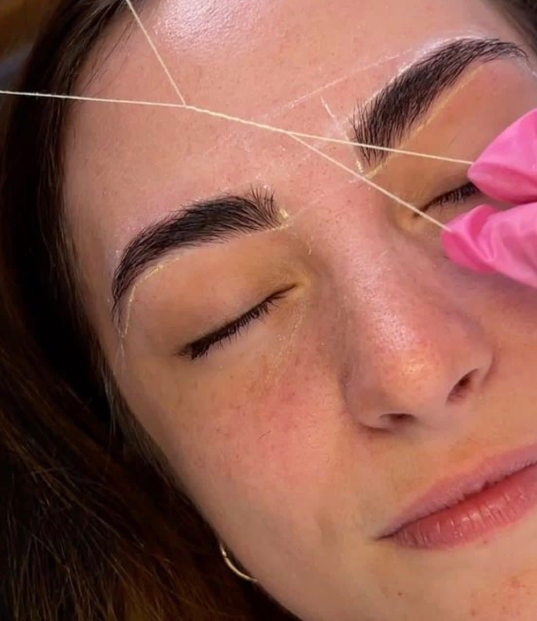 Épilation au fil sous le sourcil d’une cliente aux yeux fermés, tracé de mapping doré autour des deux sourcils, fil tendu entre des doigts gantés de rose.