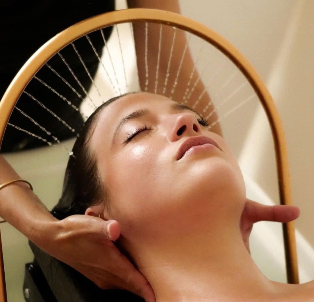 Cliente allongée, yeux fermés, sous l’arceau doré d’une douche de head spa en éventail, la nuque soutenue par les mains de la praticienne.