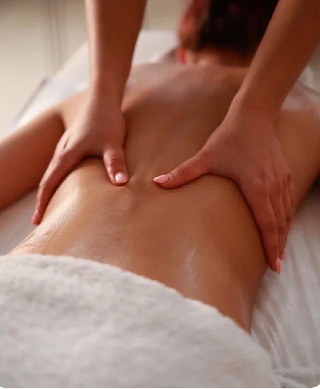 Mains glissant de part et d’autre de la colonne vertébrale sur le dos huilé d’une cliente allongée, serviette éponge à la taille.